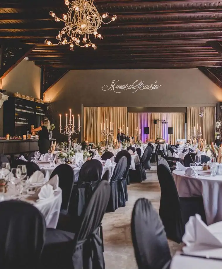 Hochzeitssaal des Mannschaftscasinos in Straubing mit schwarzem Stuhldekor und weißeingedeckten Tischen und Bühne im Hintergreund auf der die Band für Hochzeit in Straubing und Hochzeitsband in Straubing IsarSIx eine Hochzeit spielt.